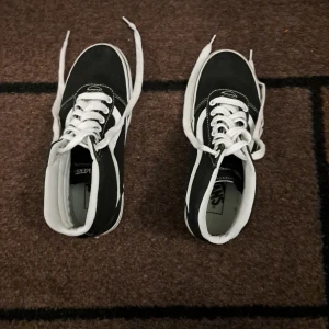 Svarta och vita Vans sneakers strl 37 - Klassiska Vans sneakers i svart och vitt med vit sula och snörning. Skorna har den ikoniska vågformen på sidan och gummisula med rutmönster undertill. Perfekta för en streetstyle-look och passar till det mesta. Storlek 37 (US 7, UK 4.5, 23.5 cm).