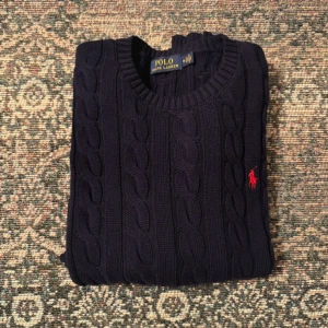 Mörkblå stickad tröja Polo Ralph Lauren - Snygg mörkblå stickad tröja från Polo Ralph Lauren med klassiskt kabelstickat mönster och röd broderad logga på bröstet. Rund halsringning och långa ärmar. Perfekt för dig som gillar stilrena och tidlösa plagg.