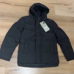 Svart dunjacka från Canada Goose M - Svart dunjacka från Canada Goose med huva och dragkedja. Jackan har klassisk patch på ärmen, två stora fickor framtill och justerbar dragsko vid huvan. Perfekt för kalla dagar och riktigt snygg streetstil. ✅Fraktar ut samma dag som du beställer ✅färg svart ✅jättesnygg vinterjacka ✅ny med tagg 