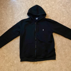 Svart hoodie från Polo Ralph Lauren - Svart hoodie från Polo Ralph Lauren med klassisk huva, dragkedja framtill och fickor. Liten vit Polo-logga broderad på bröstet. 