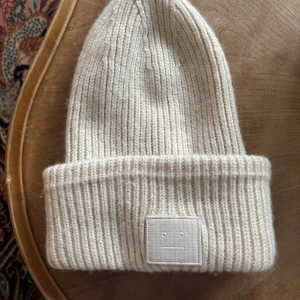 Beige ribbstickad mössa Acne Studios - Säljer en beige ribbstickad mössa från Acne Studios med deras ikoniska face-patch framtill. Mössan är i mjuk ull och har en uppvikt kant! Super bekväm och trendig. Säljer pga av att jag har köpt en ny, superbra skick❣️