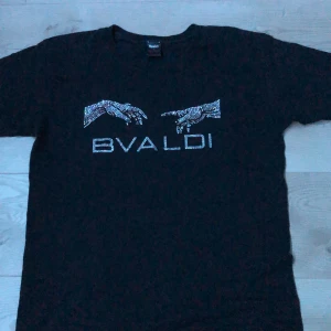 Svart t-shirt med strasstenar från Bvaldi - Svart t-shirt från Bvaldi med korta ärmar och coolt motiv av två händer och texten 'BVALDI' framtill, allt i glittrande strasstenar. Skön passform och klassisk rund hals. Perfekt för dig som gillar statement-plagg. Skick 10/10 inga defekter. Hör av dig vid frågor