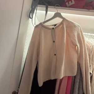 Beige kofta från Lindex med knappar - Mysig beige kofta från Lindex med rund hals och svarta knappar framtill. Tröjan har långärmad design och är tillverkad i ett fluffigt stickat material som känns skönt mot huden. Perfekt till hösten! 🍁 hör av er om frågor!