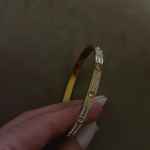 Guldigt armband med glittriga stenar - Snyggt och stilrent armband i guldfärg med glittriga stenar längs hela bandet. Armbandet har en rundad form och är dekorerat med rader av små kristaller och större stenar som ger extra bling. 