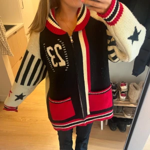 Stickad kofta från jaded London  - Säljer en oversized stickad kofta med dragkedja, stora röda fickor och coola detaljer som svarta stjärnor, ränder och siffran 23. Färgerna är svart, röd, vit och kräm. Perfekt statement-plagg med sportig vibe och bred krage.