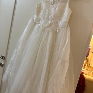 Vit långklänning för barn med spets och rosett - Superfin vit långklänning med vackra spetsdetaljer och blommiga broderier upptill. Klänningen har ett glansigt band i midjan som knyts till en rosett bak och en fluffig kjol i flera lager tyll. Perfekt för speciella tillfällen när du vill känna dig extra elegant.