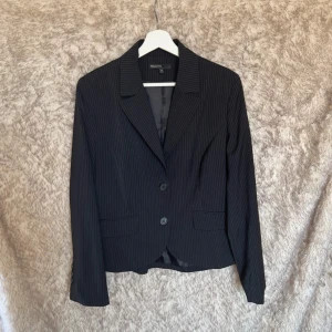 Svart randig kavaj från KappAhl - Klassisk svart kavaj med tunna vita ränder från KappAhl. Kavajen har två knappar framtill, smala slag och fickor med lock. 