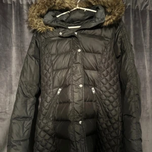 Svart dunjacka Rockandblue med fuskpäls - Svart lång dunjacka från Rockandblue med quiltade partier och snygg fuskpäls på huvan. Jackan har dragkedja, knappar, flera fickor och Rockandblue-logga på ärmen. Perfekt för kalla dagar och riktigt stilren look. I ny skick endast testad