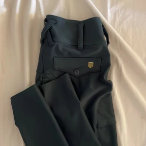 Mörkgröna ridbyxor från Maya delorez  - Mörkgröna ridbyxor från Maya delorez med hög midja och helskodd silikonmönster för extra grepp. Byxorna har praktiska fickor med lock och bälteshällor, samt guldfärgad logga på benet och bakfickan. Perfekt passform och stilren look.