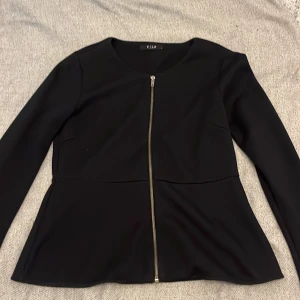 Svart kofta från Vila med dragkedja - Stilren svart kofta från Vila med lång ärm och figursydd. Koftan har en snygg silverfärgad dragkedja framtill och är tillverkad i ett mjukt, slätt material. Perfekt för dig som gillar en clean look med edge.