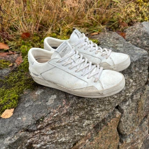 Vita sneakers från Crime London - Snygga vita sneakers från Crime London med klassisk låg profil och rund tå. Skorna har snörning och är tillverkade i skinn med diskreta detaljer och logga på hälen. Perfekta för dig som gillar stilrena och enkla sneakers med urban känsla.