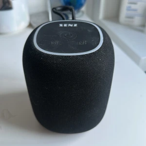 Senz SpodBK Portable Speaker - Senz SpodBK är en portabel högtalare med snygg svart design. Den har USB- och AUX-anslutning, tydliga knappar för styrning och levereras med tillhörande kablar. Högtalaren är i gott skick med mindre yttre damm, perfekt för musik på språng eller hemma. Ny pris: 499kr 