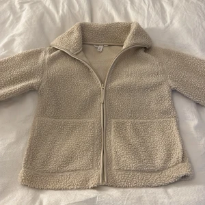 Beige teddyjacka från H&M - Mysig beige teddyjacka från H&M med dragkedja framtill och två stora fickor. Jackan har hög krage och är tillverkad i mjuk fleece som håller dig varm under kyliga dagar. Perfekt för dig som gillar en chill och avslappnad stil.