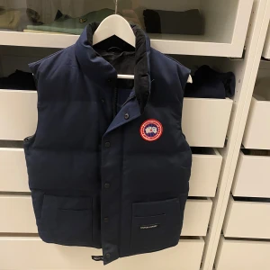 Mörkblå dunväst från Canada Goose - Snygg mörkblå dunväst från Canada Goose med klassisk logga på bröstet. Västen har hög krage, två stora fickor framtill och stängs med både dragkedja och knappar. Perfekt för lager-på-lager och riktigt varm under kyliga dagar.