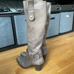 Bruna stövlar med klack från Vai vei - Snygga höga stövlar i brunt skinn med rund tå och stabil klack. Stövlarna har dekorativa remmar med metallknappar och dragkedja på insidan för enkel påtagning. Perfekt till jeans eller kjol för en cool look.