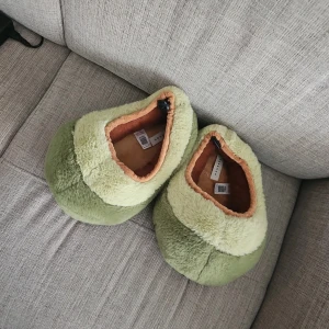 Gröna fluffiga avokado tofflor från Topshop - Mysiga tofflor från Topshop i två nyanser av grönt med beige insida. Utsidan är fluffig och mjuk, insidan har en varm plyschkänsla. Rund tå och platt sula med halkskydd. Perfekta för chill hemma och håller fötterna varma under kalla dagar. Storlek 39-40