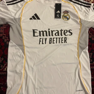 Real Madrid vit fotbollströja Adidas - Snygg vit Real Madrid fotbollströja från Adidas med svarta detaljer och gula linjer längs sidorna. Tröjan har korta ärmar, klubbmärke på bröstet och Emirates Fly Better-tryck. Tillverkad i lätt och ventilerande polyester med AEROREADY-teknologi.