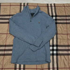 Blå stickad tröja Polo Ralph Lauren - Snygg blå stickad tröja från Polo Ralph Lauren med halv dragkedja och klassisk broderad logga på bröstet. Tröjan har lång ärm, ribbade muddar och hög krage. Perfekt för lager-på-lager och chill stil.