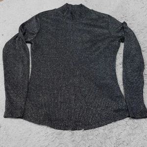 Glittrig svart polotopp från Lindex - Säljer en snygg svart polotopp från Lindex i storlek S. Toppen har ett glittrigt material och lång ärm, med en hög krage som ger en cool och trendig look. Perfekt för dig som gillar att sticka ut med lite extra glitter i outfiten.