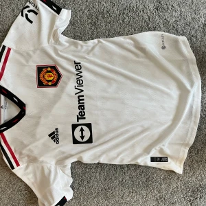 Manchester United vit bortatröja Adidas - Säljer en vit Manchester United bortatröja från Adidas med svart och röd detalj vid krage och ärmslut. Tröjan har klubbmärke på bröstet, TeamViewer-tryck och Adidas-logga. Materialet är lätt och andas, perfekt för match eller träning.