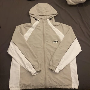 Beige och vit vindjacka från Corteiz - Snygg beige och vit vindjacka från Corteiz med huva och hel dragkedja. Jackan har svarta detaljer, elastiska muddar och meshfoder. Broderad logga på bröstet och tryck på ryggen. Perfekt för dig som gillar streetwear och sportig stil.