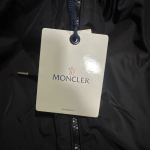 Svart vindjacka från Moncler med huva - Svart vindjacka från Moncler med klassisk logotyp på ärmen och dragkedja framtill. Jackan har huva med röd, vit och blå randig detalj och är tillverkad i lätt polyester. Perfekt för dig som vill ha en stilren och sportig look.