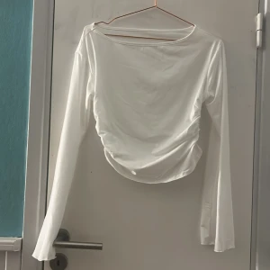 Vit långärmad omlottopp - Säljer en vit långärmad topp med omlottdetalj och lätt rynkning i midjan med vida armar. Toppens material är mjukt och har en slät yta, perfekt för dig som gillar stilrena och enkla plagg med en twist. Passar dig som vill ha en clean look med lite extra. Testad en gång, liten i storlek