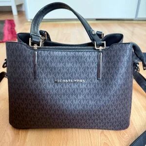 Michael Kors brun handväska med logomönster - Snygg handväska från Michael Kors i mörkbrunt med MK-logomönster över hela väskan. Väskan har svarta handtag, avtagbar axelrem och dragkedja upptill. Tillverkad i skinnliknande material och har flera fack inuti för smart organisering.