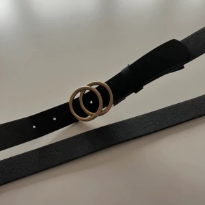 Svart bälte med dubbel ring-spänne - Stilrent svart bälte med ett snyggt spänne i form av två guldfärgade ringar. Bältet är slätt och har fem hål för justering. Materialet är skinnimitation och perfekt för att lyfta din outfit med en cool detalj. 85 cm (33 inch)