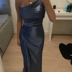 Marinblå långklänning i satin - Säljer en elegant marinblå långklänning i glansigt satinmaterial. Klänningen har oneshoulder-design, figurnära passform och en snygg slits framtill. Draperingarna ger en lyxig känsla och den är perfekt för bal eller fest. Supersnygg och stilren look! Använd endast en kväll