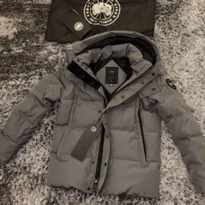 Canada goose jacka  - Snygg mörkgrå Canada Goose-jacka i storlek M med luva och svart loga på armen. Jackan har både dragkedja och knappar framtill för extra skydd mot kylan. Stilren och varm – perfekt för kalla dagar när du vill hålla stilen och hålla dig bekväm. Svarar alltid alla meddelanden direkt och fraktar alltid samma dag som köp:)