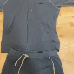 Boss ser - Snygg mörkgrå set från BOSS med dragkedja framtill och diskret logga på bröstet. Tröjan har långa ärmar, fickor på sidorna och kontrastdetaljer längs axlarna. Perfekt för chill eller träning, riktigt skön och stilren.