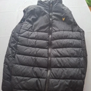 Svart väst från Lyle & Scott Junior - Svart quiltad väst från Lyle & Scott Junior med gul broderad logga på bröstet. Västen har hög krage och dragkedja framtill. Perfekt för lager-på-lager och har en stilren, sportig look som funkar till många outfits.