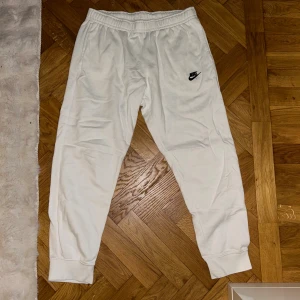Nike Mjukisbyxor  - Säljer ett par vita mjukisbyxor från Nike med klassisk svart logga på vänster lår. Byxorna har resår i midjan och muddar vid bensluten för en skön och avslappnad passform. Perfekta för chill eller träning.