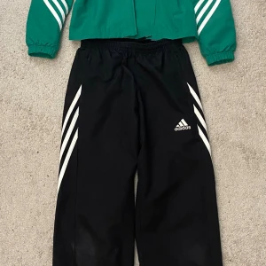 Svarta adidas set med vita ränder - Svart set från Adidas med klassiska vita ränder längs sidorna och Adidas-logga på låret. Byxorna har elastisk midja och är tillverkade i ett lätt syntetmaterial som passar perfekt för sport och träning.