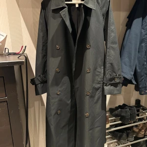 Svart trenchcoat från H&M XS - Säljer en svart trenchcoat från H&M i storlek XS men sitter som en s. Jackan har dubbelknäppning med silvriga knappar, klassisk krage och bälte i midjan samt vid ärmsluten. Lång modell med stilren look, perfekt för dig som gillar minimalistisk stil.
