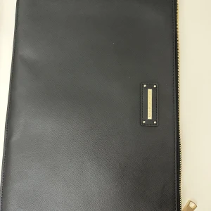 Svart datorväska från Ideal of Sweden - Svart datorväska från Ideal of Sweden med guldfärgade dragkedjor och metallplatta med logga. Väskan har en slät, lätt texturerad yta och ett smalt, rektangulärt format. Perfekt för att bära din laptop och tillbehör med snygg vibe. Mått 39 x 27cm