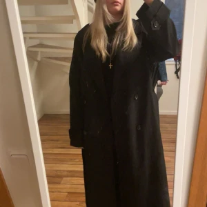 Svart trenchcoat från NA-KD, stl 38 - Säljer en svart trenchcoat från NA-KD i storlek 38. Jackan har klassisk dubbelknäppning, bred krage, bälte i midjan och snygga detaljer vid ärmslut och axlar. Perfekt för dig som gillar en oversized och stilren look.