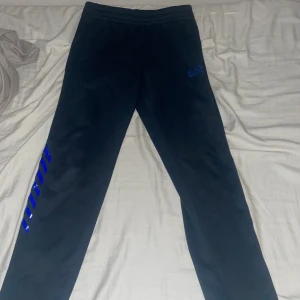 Svarta EA7 mjukisbyxor med blå detaljer - Svarta mjukisbyxor från EA7 Emporio Armani med blå logga på framsidan och blå ränder längs benen. Byxorna har elastisk midja med snörning och är gjorda i mjukt material som är perfekt för chill eller träning. Snygg och sportig look med slim passform. Storlek xs men passar S. Använt fåtal gånger.