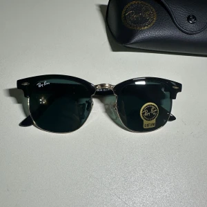 Svarta solglasögon från Ray-Ban - Snygga Ray-Ban Clubmaster solglasögon i klassisk svart färg. De har en stilren design med mörka glas och en elegant båge. Perfekta för en cool och tidlös look.