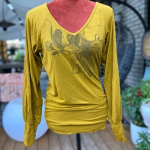 Gul långärmad tröja Komodo 36 - Senapsgul långärmad tröja med V-ringning och ett tryck framtill. Tröjan har en åtsittande passform och muddar vid ärmsluten. Den har även en detalj med dragsko baktill. Storlek: 1 ﻿Bomull Längd: 70 cm Bystmått: 49 cm   Inga bilder när den sitter på mig! Katter finns i hemmet!