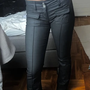 Svarta byxor med fickdetaljer - Snygga svarta byxor med raka ben och markerad söm framtill. Byxorna har två stora fickor med lock framtill och stängs med knapp och dragkedja. Materialet har en lätt glansig finish som ger en cool look.