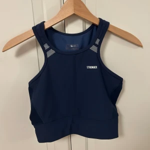 Marinblå linnetopp från Stronger - Snygg marinblå linnetopp från Stronger med  design och vita detaljer vid axlarna. Hög halsringning, och rygg. Har ett stretchigt syntetmaterial som är perfekt för träning. Loggan syns både fram och bak. Nästintill oanvänd men loggan inuti är inte jättesynlig. Köpt för 549 kr. 