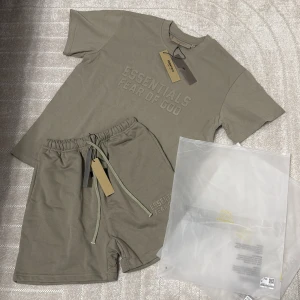 Olivgrön t-shirt och shorts Essentials Fear of God - Snygg beige t-shirt från Essentials Fear of God med tryckt logga framtill. Klassisk rund halsringning och kort ärm. Tillverkad i mjuk bomull för en skön känsla. Perfekt för en clean och avslappnad stil.