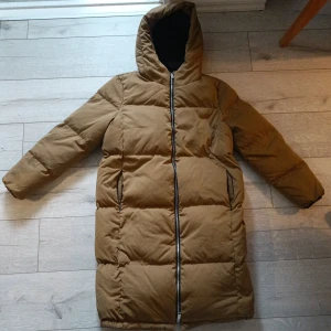 Brun lång pufferjacka från Y.A.S XS - Säljer en brun lång pufferjacka från Y.A.S i storlek XS. Jackan har huva, hel dragkedja framtill och två fickor med dragkedja. Perfekt för kalla dagar med sin vadderade design och stilrena look.