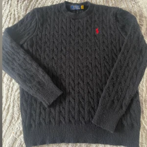 Svart stickad tröja Polo Ralph Lauren - Svart kabelstickad tröja från Polo Ralph Lauren med rund hals och lång ärm. Tröjan har ett rött broderat Polo-logotyp på bröstet och ribbade muddar vid ärmslut och nederkant. Perfekt för dig som gillar klassisk stil med en twist. Storleken är L men passar som M-S
