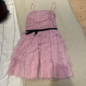 Cool rosa volangklänning med rosett - Cool rosa kortklänning med svarta prickar upptill och flera lager volanger i kjolen. Klänningen har smala axelband och ett svart band med rosett i midjan. Jättebra skick💕