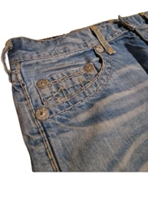 True Religion wide jeans i ljusblå - Säljer ett par ljusblå jeans från True Religion med breda ben och klassiska kontrastsömmar. Jeansen har detaljerade fickor med knappar och två stora True Religion-märken på bak fickorna. Materialet är robust denim och passformen är loose med wide stil.