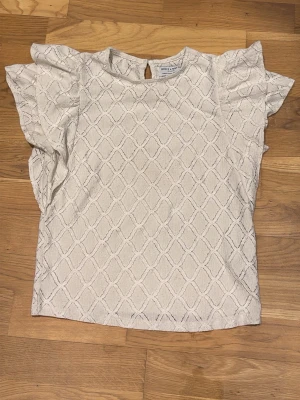 Vit blus med volangärmar Holly & White - Säljer en vit blus från Holly & White med snyggt strukturmönster och volangdetaljer på axlarna. Blusen har rund hals och knapp i nacken. Perfekt för dig som gillar romantiska detaljer och en ljus look.