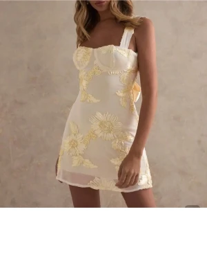 Gul kortklänning med blommor och rosett - Supersöt kort klänning i gul mesh med stora broderade blommor i ljusgult. Klänningen har breda axelband, öppen rygg med knytband i nacken och en stor rosett bak. Figurnära passform och slits i sidan. Perfekt för sommarens alla tillfällen! Nypris: 350